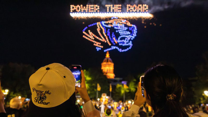 Two students holding smart phones photograph the drone show above the dome of Jesse Hall as drones spell out Power the Roar