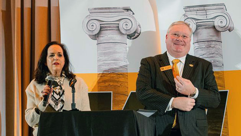 Lynn Ann Vogel and Paul Vogel speak in front of banners displaying columns.