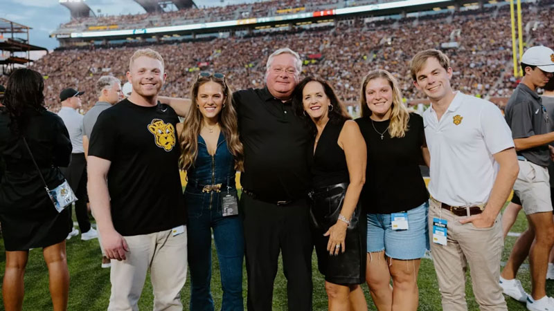Paul and Lynn Vogel stand with their family outside the end zone of Faurot Field with a packed stadium behind them.