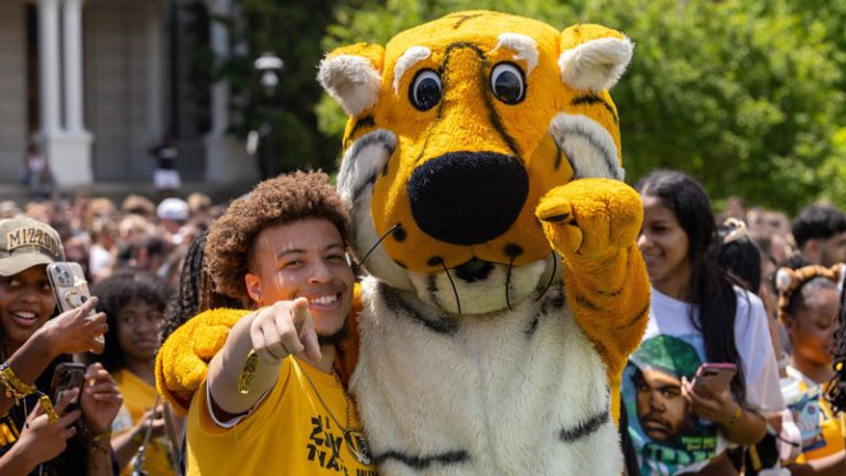 A Mizzou student and Truman the mascot point at the camera while surrounded by a croud of students.