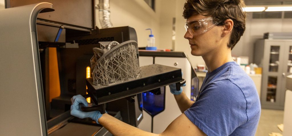 Student wearing safety glasses and gloves removes a newly printed metal component from a 3D printer inside a university engineering lab.