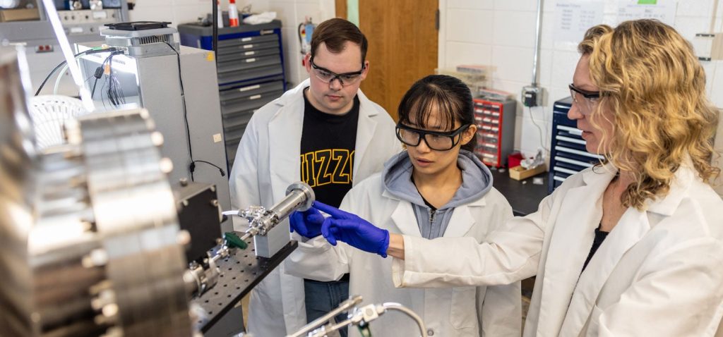 Three researchers wearing lab coats and safety glasses examine laboratory equipment in a campus lab setting.