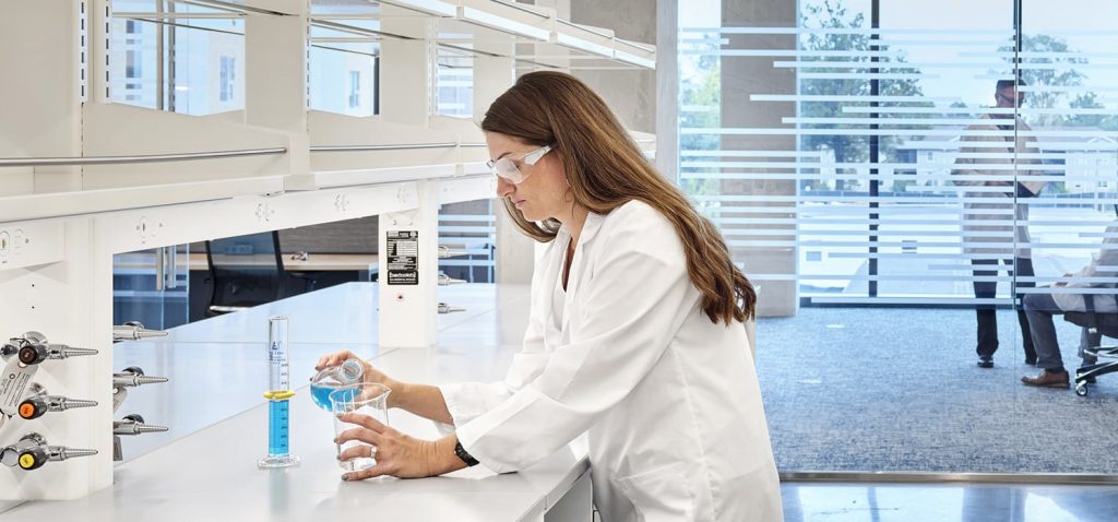 Scientist in a white lab coat and safety glasses works at a laboratory bench, with colleagues visible in a glass-walled office behind her.