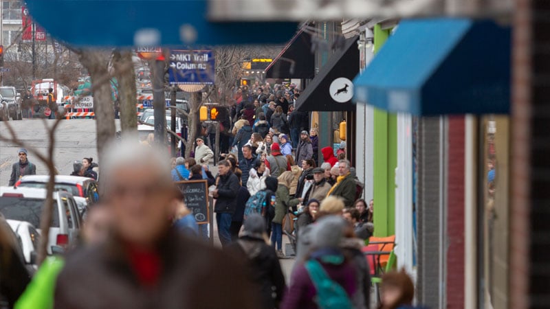 Crowds walk down the sidewalk in front of storefronts in downtown Columbia, Missouri.